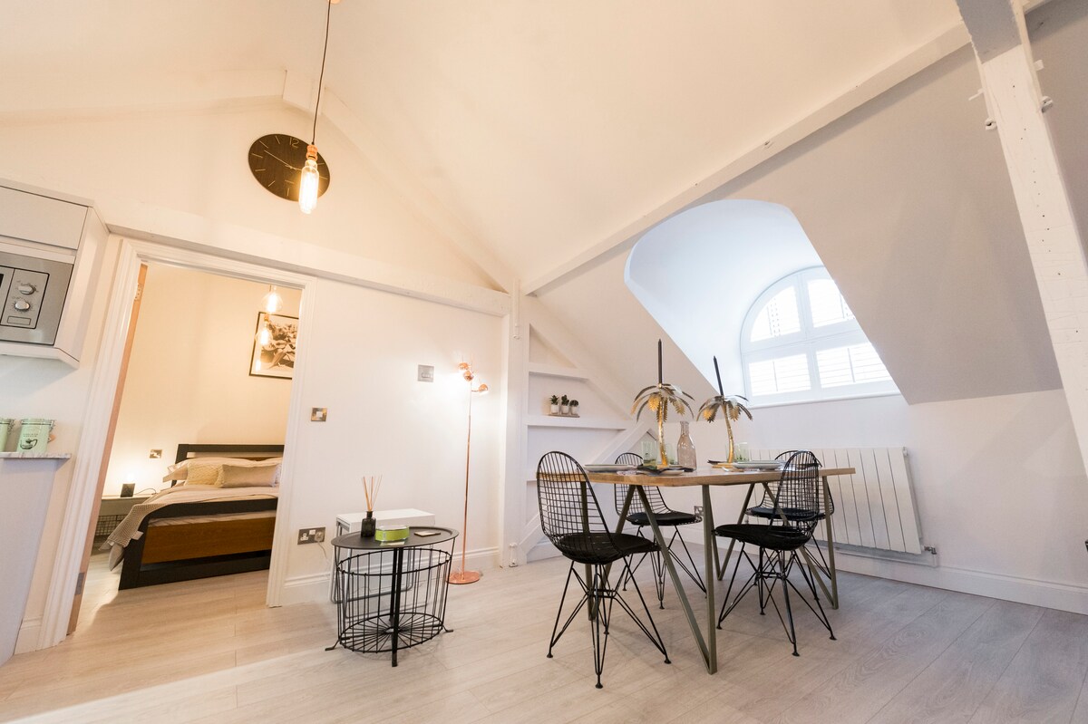 A well-lit dining area features a sleek table with two wire chairs and a small side table. Beyond the arch of the wall, a cozy bedroom is partially visible, with a spacious bed and decorative artwork. Natural light filters through a traditional window.