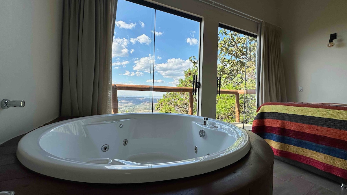A spacious hot tub is positioned beside large glass doors that open to a scenic view of the landscape. The room features a colorful striped blanket on the bed, with natural light illuminating the space.