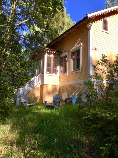 The exterior of a traditional nineteenth-century Swedish country house is displayed, featuring yellow wooden siding and large windows. A staircase leads to a small porch, framed by greenery. Two wooden chairs and a bicycle can be seen in the foreground, adding to the serene garden setting.