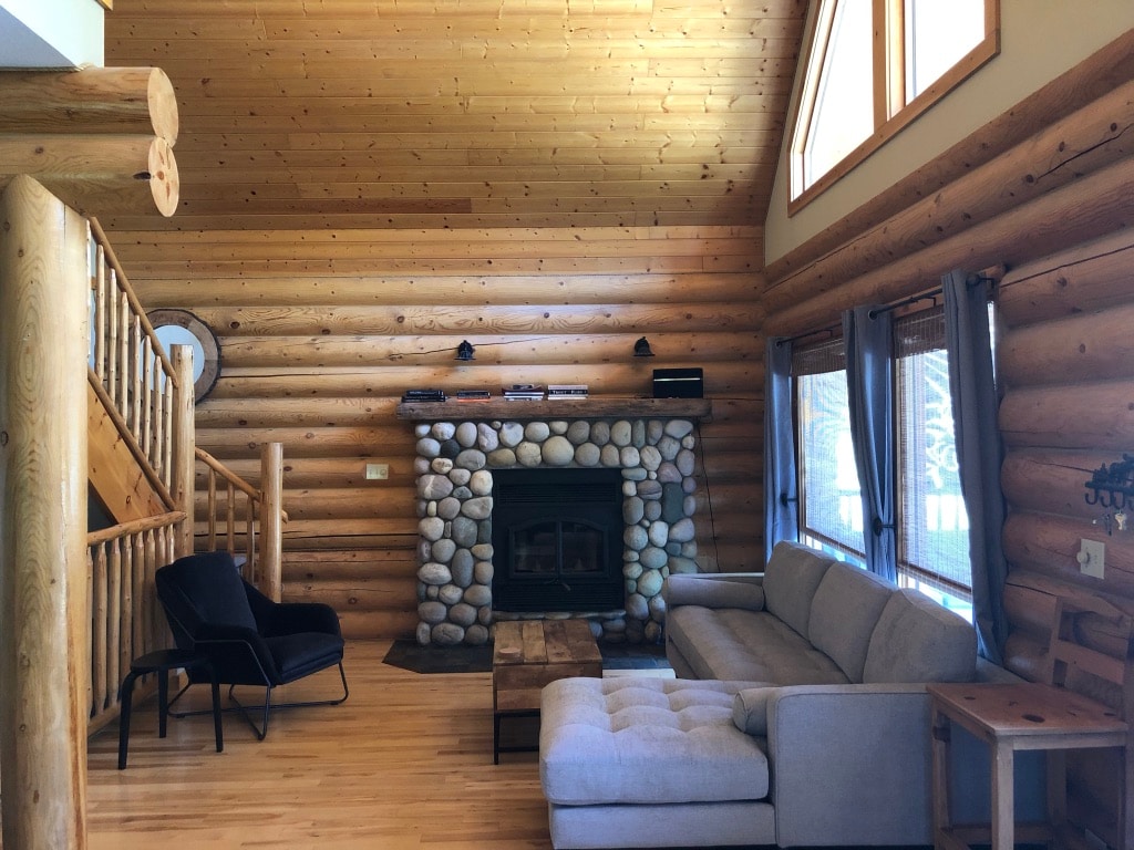 A spacious living area features a comfortable sectional sofa and two armchairs. A stone fireplace serves as the focal point, flanked by large windows that allow natural light to illuminate the wooden walls and ceiling, enhancing the cabin's rustic charm.