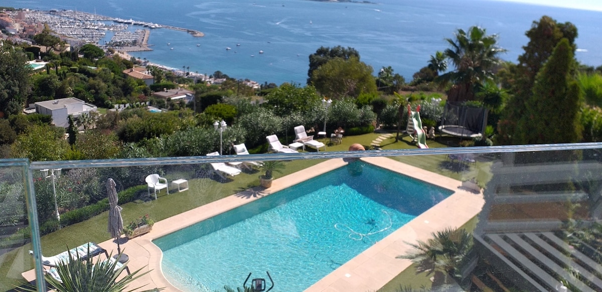 A sparkling swimming pool is featured within a manicured garden, surrounded by lounge chairs and trees. A panoramic view of the sea and coastline is visible in the background, showcasing the natural beauty of the surroundings.