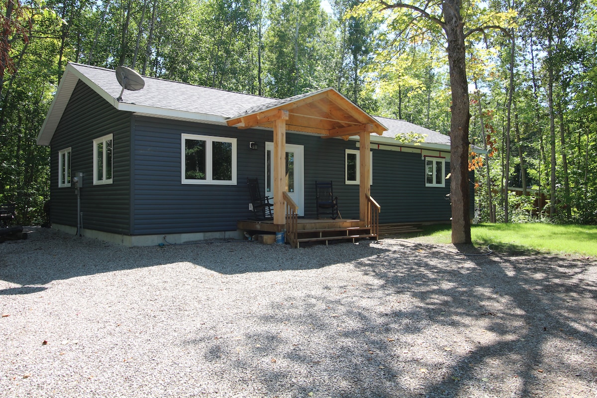 A well-structured house is set within a wooded area, surrounded by mature trees. The front porch features a wooden overhang and is flanked by rocking chairs. A gravel driveway leads to the entrance, providing a rustic yet inviting approach.
