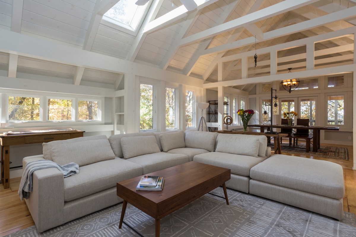 A spacious living area is showcased, featuring a large sectional sofa and a low wooden coffee table. Natural light floods in through multiple windows, highlighting the rustic wood beams and open floor plan that connect to a dining space in the background.