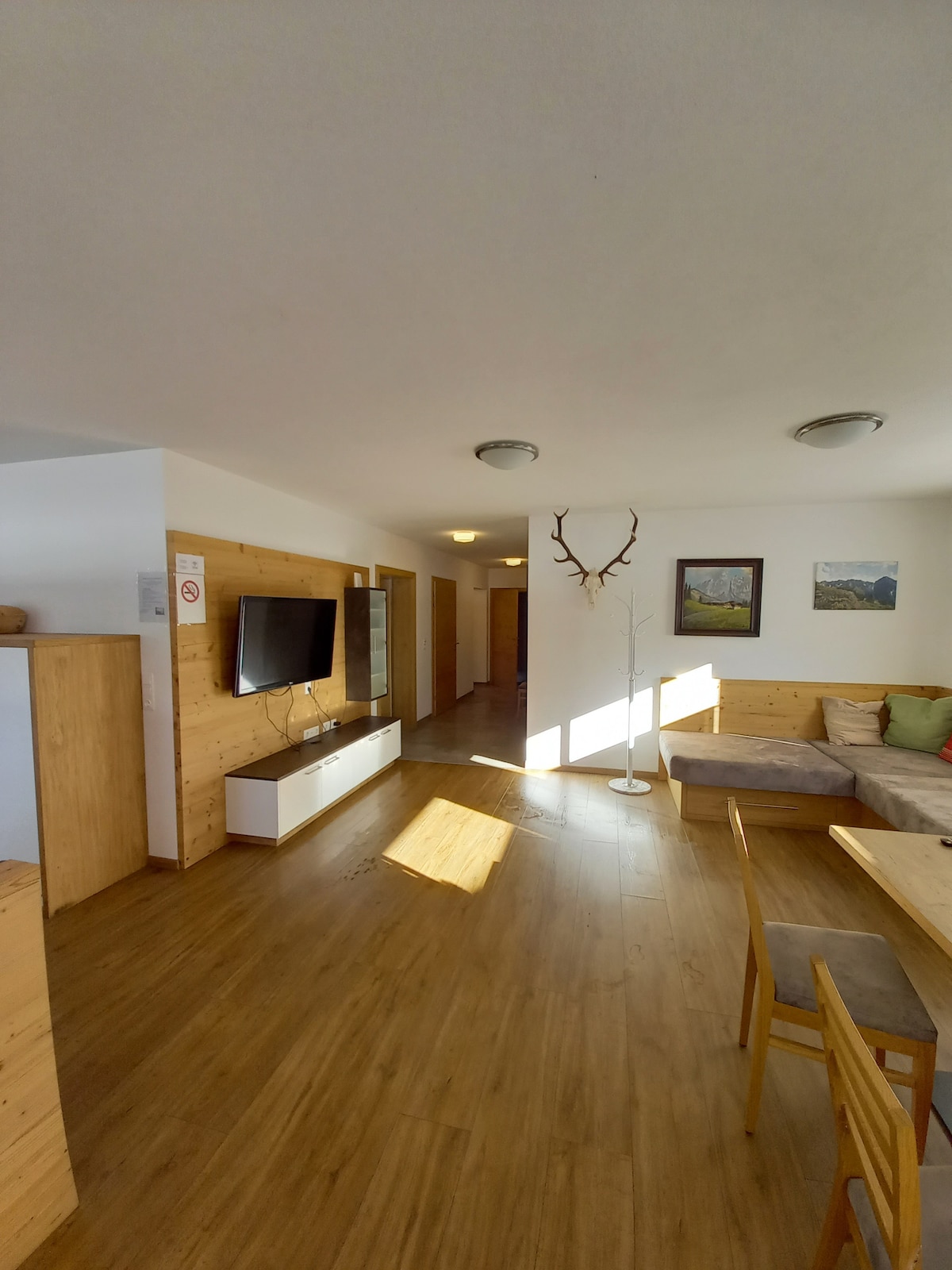 A spacious living area is featured with warm wooden accents and natural light streaming in from multiple sources. A flat-screen TV is mounted on a light-colored cabinet, while a dining table and chairs are positioned nearby, inviting moments of relaxation and connection.