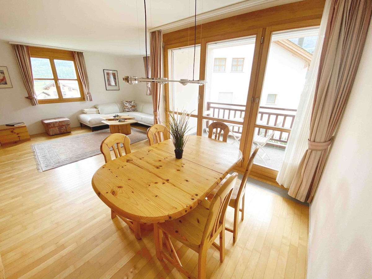 A spacious dining area features a wooden table surrounded by six chairs, with natural light pouring in through large windows. A cozy living area is visible in the background, and warm wooden accents enhance the inviting atmosphere.