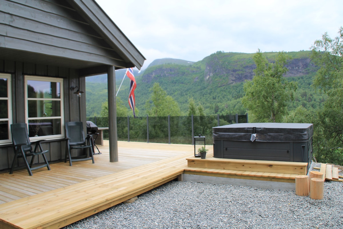 A wooden deck extends from the cabin, featuring a hot tub and a seating area with two chairs. The landscape showcases rolling green hills under a cloudy sky, with a flagpole displaying the national flag nearby.