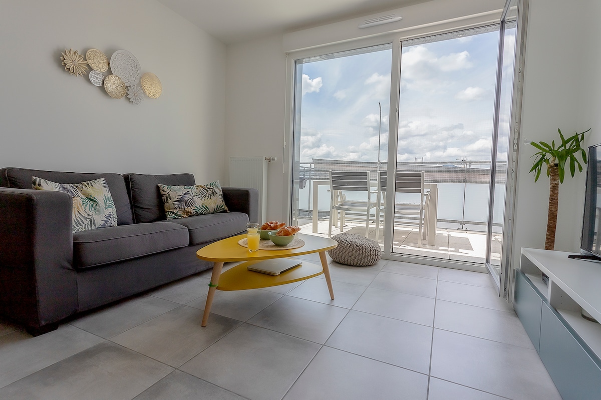 A bright living area features a dark gray sofa adorned with decorative throw pillows. A yellow coffee table sits in front, topped with a bowl of fruit. Large glass doors open onto a balcony, allowing natural light to fill the space and offering views of the outdoors.