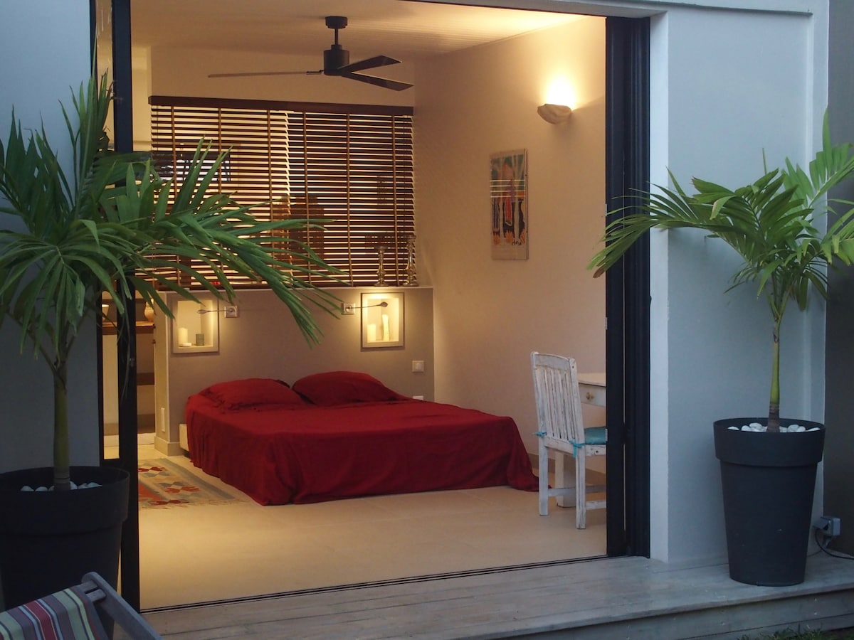 A spacious bedroom is visible, featuring a large bed with a red coverlet. Two bedside lamps are mounted on the wall, and a ceiling fan is positioned overhead. Light filters in through partially open wooden blinds, enhancing the calming ambiance of the space.