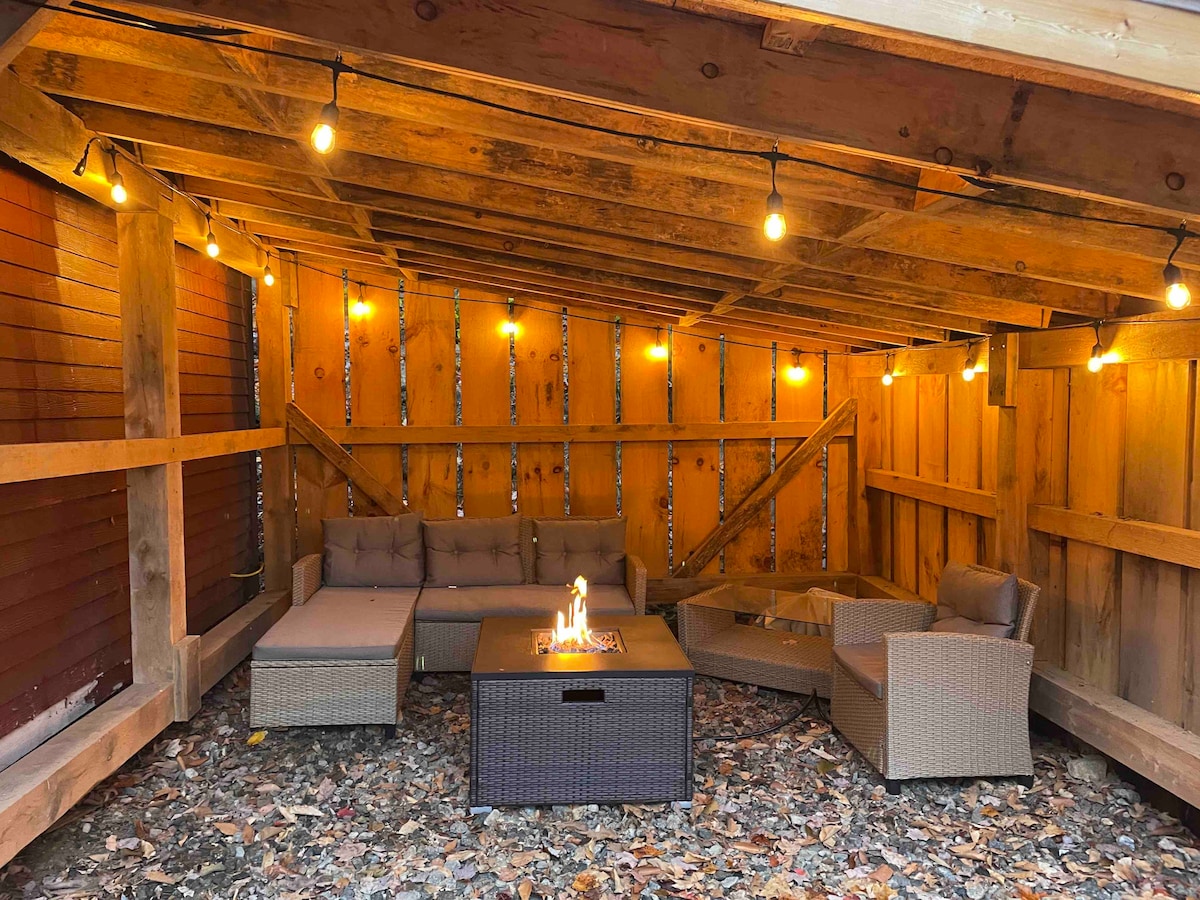 A covered outdoor patio is illuminated by subtle string lights, creating a cozy environment. Comfortable seating is arranged around a gas fire pit, with a glass tabletop positioned centrally. The ground is adorned with a layer of fallen leaves, enhancing the natural ambiance.