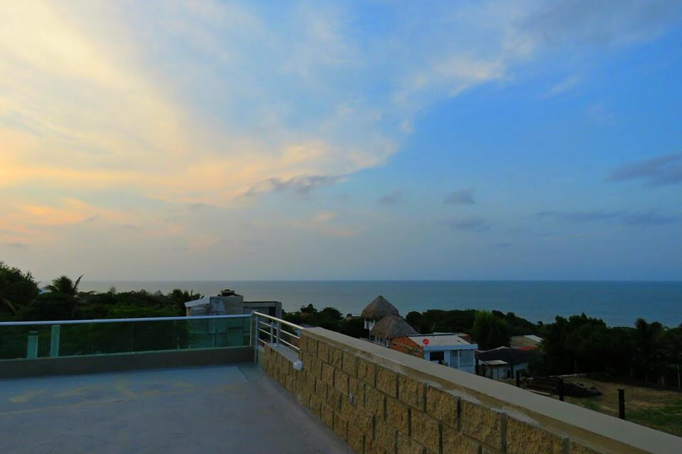 A panoramic view of the sea is presented from the terrace, with the horizon showcasing a blend of soft blues and warm hues. The sky features scattered clouds, while the surrounding landscape includes greenery and distant buildings, enhancing the tranquil atmosphere.