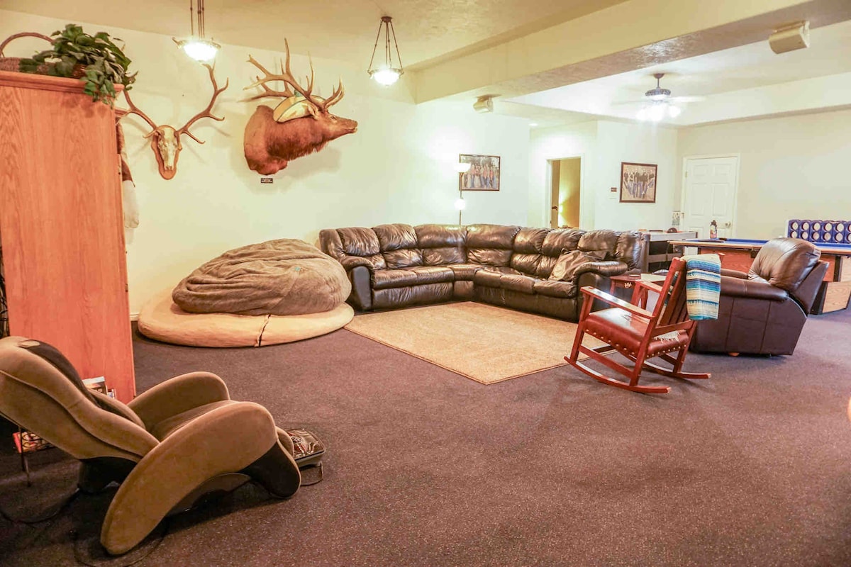 A spacious living area features a large sectional leather sofa arranged around a central rug. Decorated with mounted animal heads, the room is softly lit with ceiling fans overhead. A rocking chair and a unique bean bag chair add to the relaxing atmosphere.