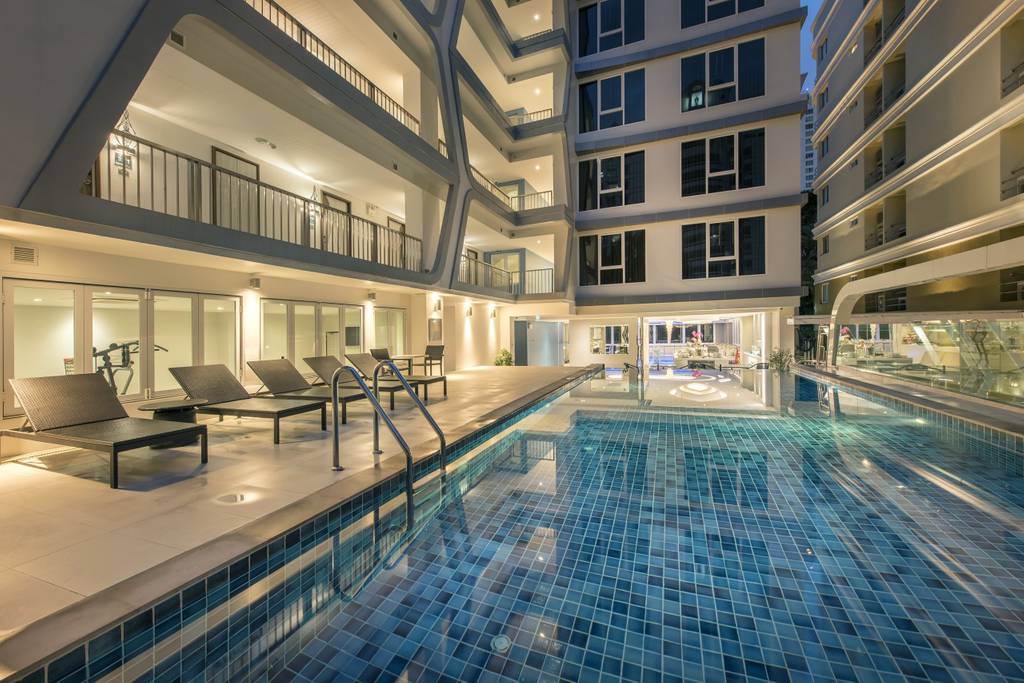 A tranquil swimming pool is surrounded by a modern building structure, with lounge chairs positioned alongside. Indoor spaces are visible through large glass doors, reflecting a serene evening ambiance. Soft blue tiles line the pool, enhancing the calm atmosphere of the area.
