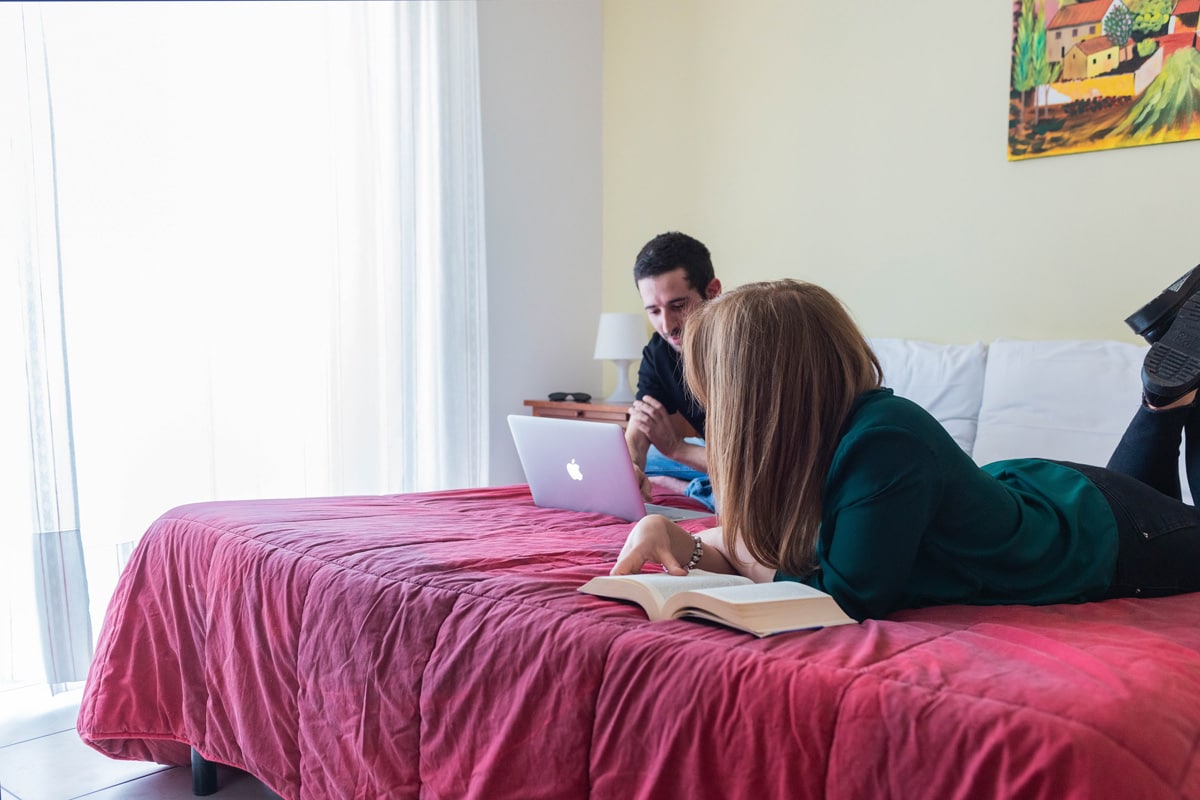 A spacious bed with a rich red cover is centrally placed in the room. A woman is seen lounging, reading a book, while a man sits nearby using a laptop. Natural light floods in from large glass doors that lead to a balcony.