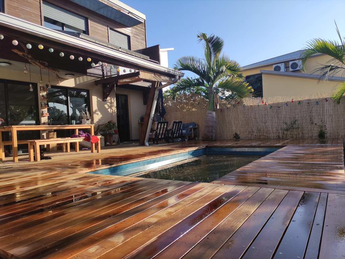 A wooden deck surrounds a clear swimming pool, inviting relaxation. The area is adorned with potted plants and a dining table. Comfortable seating options are visible under a shaded structure, with sunlight streaming in, creating a warm atmosphere.