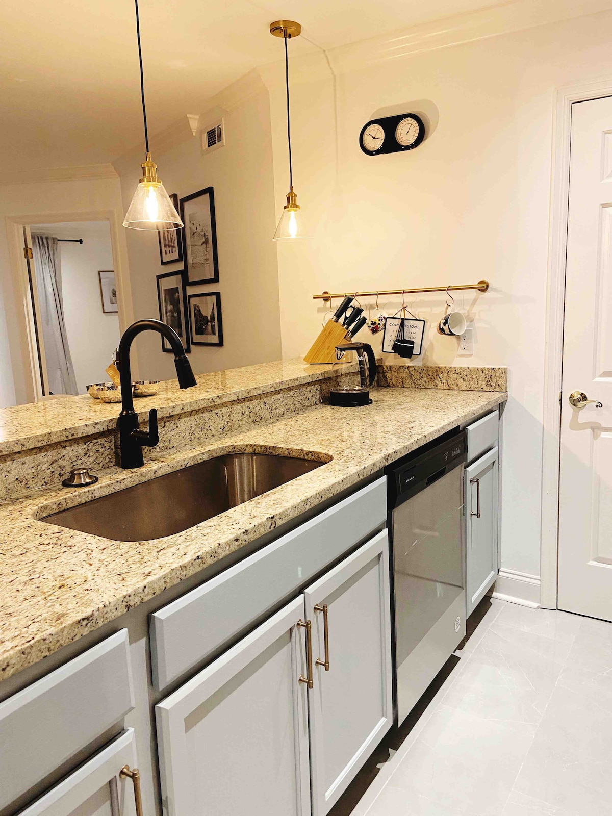 A modern kitchen area features sleek granite countertops and a stainless steel sink. Under-cabinet lighting highlights the space, while a decorative clock and hanging utensils add functional charm. Nearby, a dishwasher is visible, complementing the stylish cabinetry.