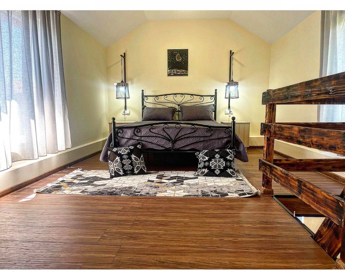 A charming sleeping area features a wrought-iron bed dressed in dark bedding, flanked by wall-mounted lamps. A textured rug lies beneath the bed, and large windows adorned with light curtains allow natural light to fill the space.