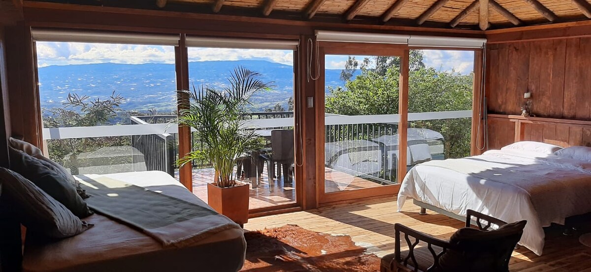 A cozy interior features two spacious beds beside large glass doors, which provide a stunning view of the valley. Natural light fills the room, highlighting the wooden beams of the ceiling and the earthy tones of the decor. A potted plant adds a touch of greenery.