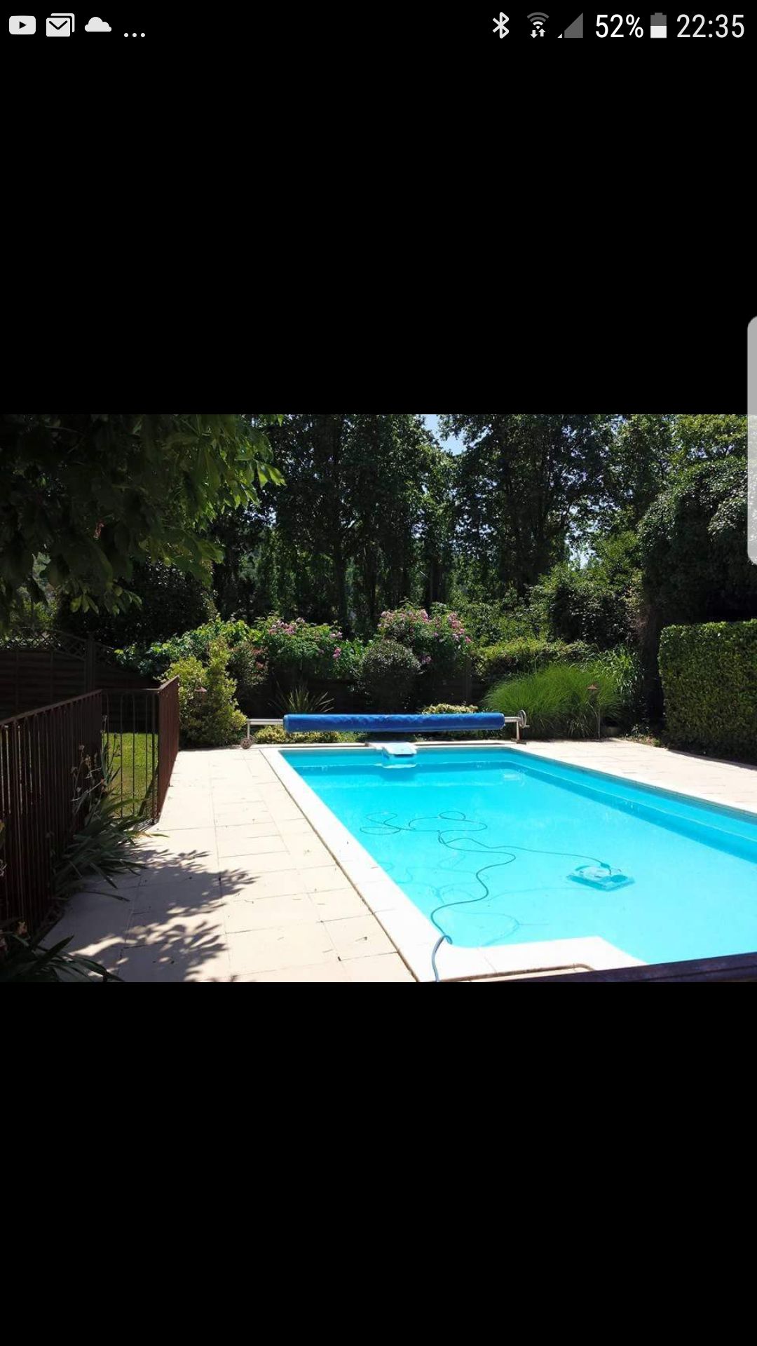 A private outdoor swimming pool is surrounded by lush greenery and flowering plants. Sunlight reflects off the clear water, and a shaded area is indicated by a blue cover at one end. A tiled deck borders the pool, providing a space for relaxation.