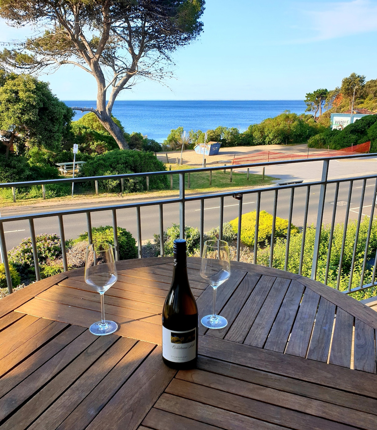 A wooden table is set with two empty wine glasses and a bottle of wine, positioned on a balcony. In the background, sweeping views of the bay and coastal greenery are visible, complemented by a clear blue sky.