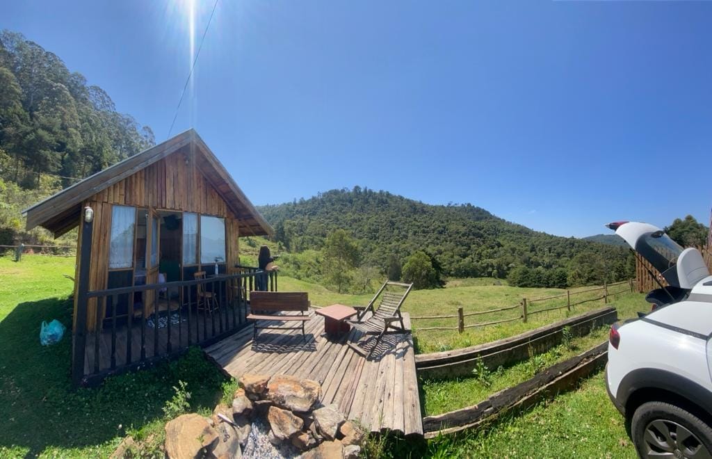 A wooden cabin with a deck overlooks a green landscape, featuring a mountain in the background. The deck includes a seating area and a lounge chair. A car is parked nearby, and the sky is clear with bright sunlight illuminating the scene.