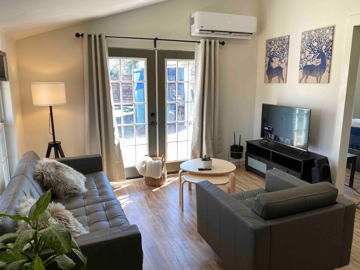 A living area features a modern layout with two comfortable sofas, a round coffee table, and a flat-screen television. Natural light fills the space through glass doors leading to an outdoor area. Decorative plants are positioned nearby, adding a touch of greenery.