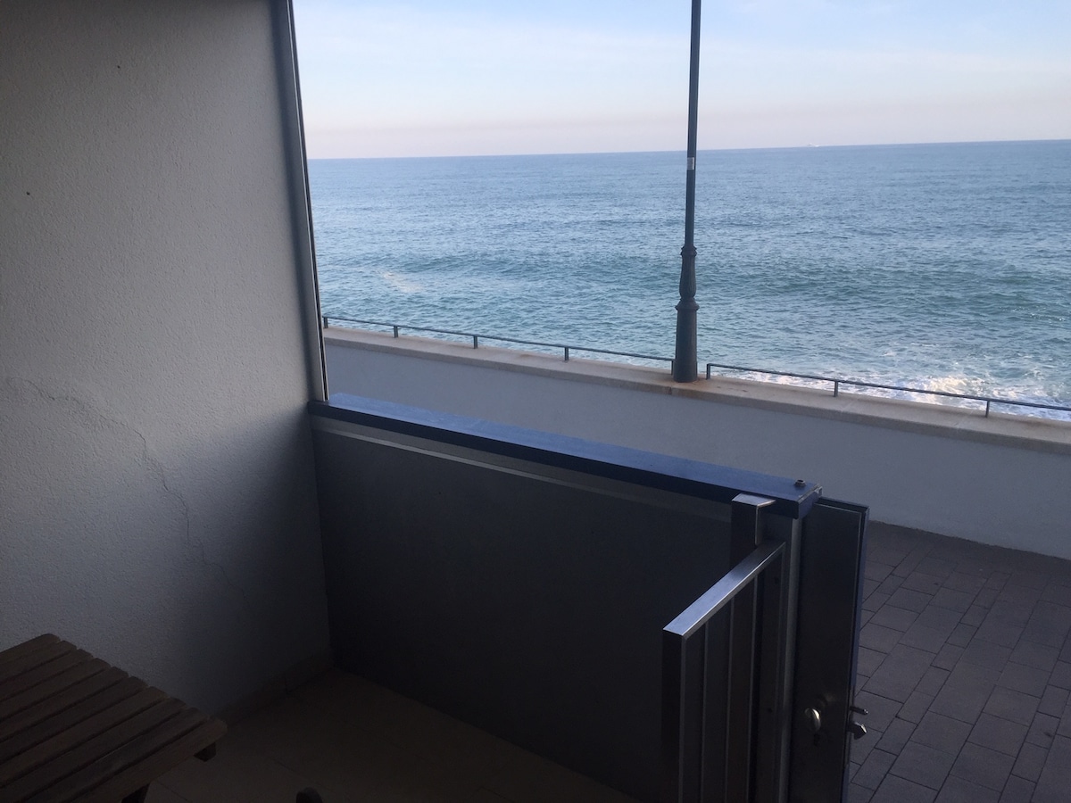 A balcony is shown with a clear view of the ocean, featuring calm blue waters meeting the horizon. A wooden table is positioned against the wall, and natural light filters through the open space, contributing to a serene coastal atmosphere.