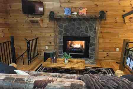 A cozy living area features a stone fireplace set against warm wooden walls, with a comfortable sofa and a rustic coffee table positioned nearby. A flat-screen TV is mounted above the fireplace, completing the inviting atmosphere.