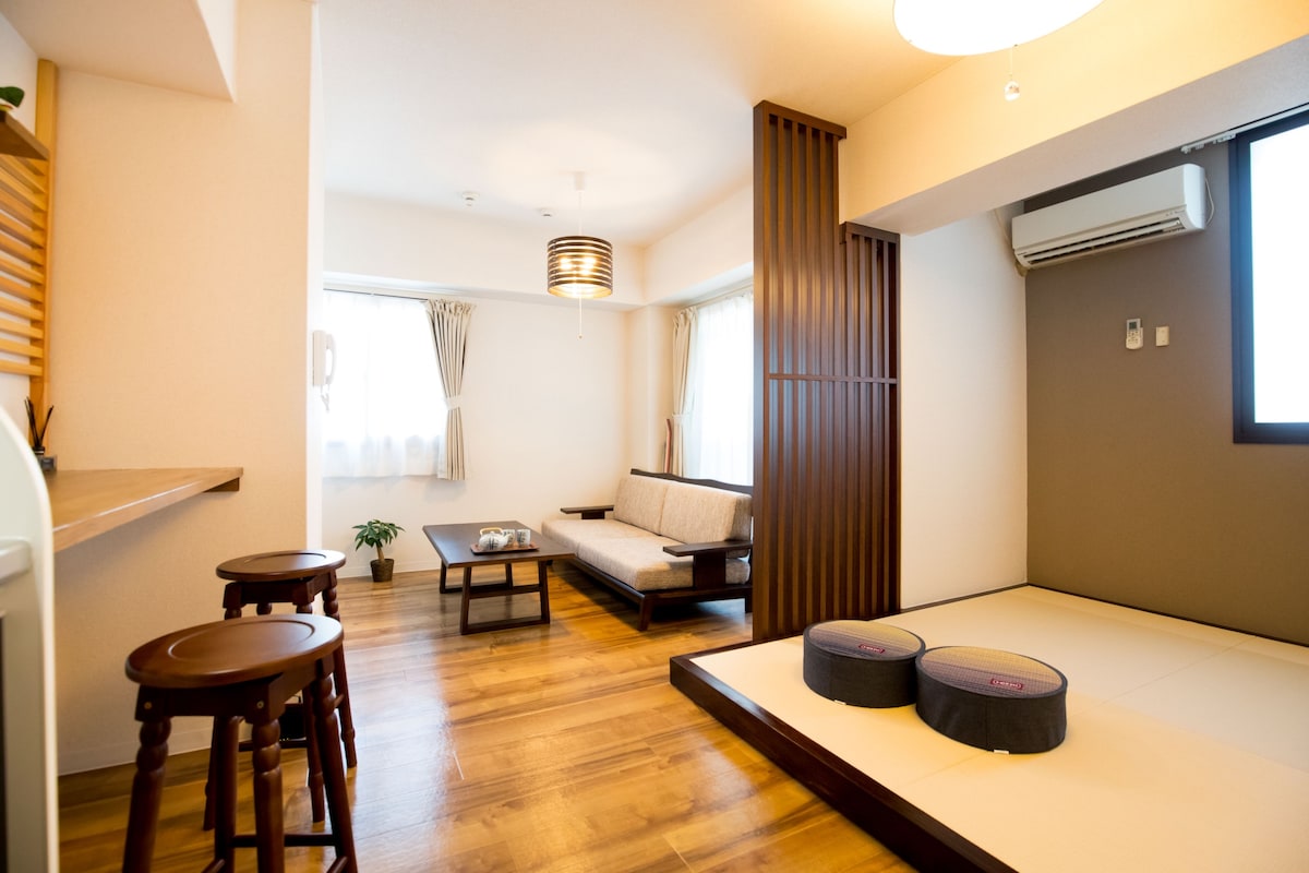 A living area features a light wood floor with tatami mats in one corner. A low table is positioned with two cushions nearby. A sofa is set against a wall, accompanied by a window with sheer curtains allowing natural light. Air conditioning is mounted above.