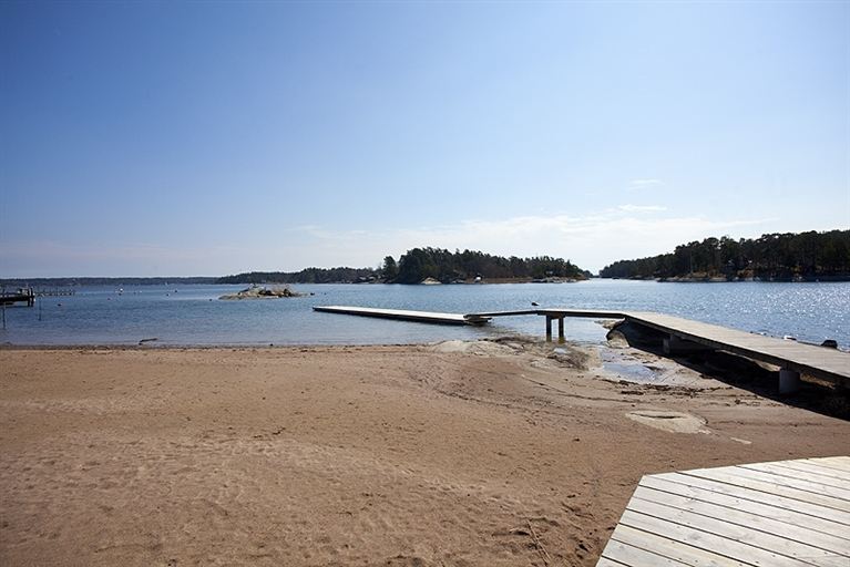 A sandy shoreline is seen leading to a calm body of water, with wooden docks extending into the lake. The scene is illuminated by bright sunlight, with distant trees lining the horizon under a clear blue sky.