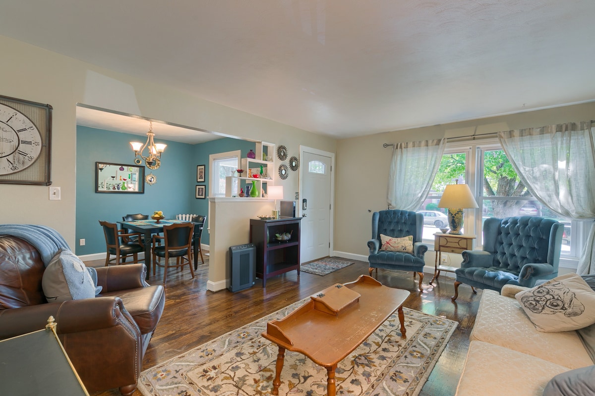 A cozy living area features a mix of comfortable seating, including two blue armchairs and a leather sofa. A patterned area rug sits on polished wooden flooring. A dining area is visible in the background, illuminated by natural light from the window.