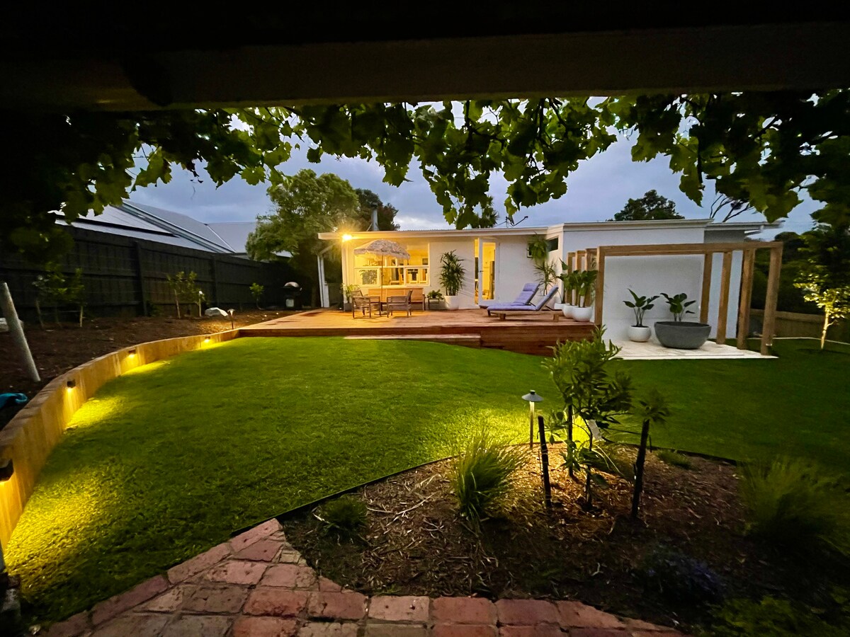 The serene backyard is illuminated by soft lighting, featuring a well-maintained lawn and garden beds. The house, with a wooden deck, is visible in the background, showcasing large windows and outdoor seating. Lush greenery surrounds the area, enhancing the tranquil ambiance.