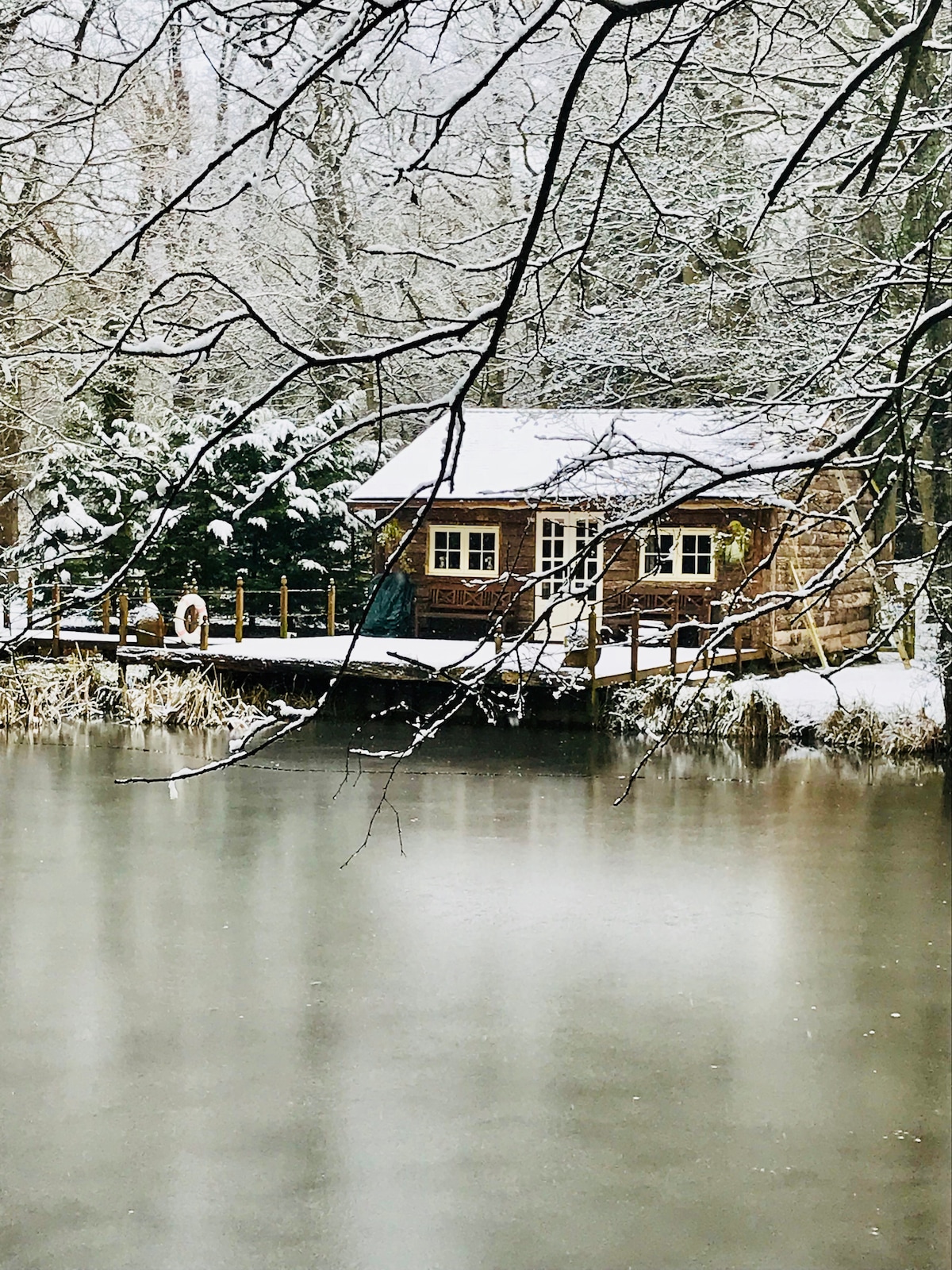 A charming holiday cabin is set against a serene, snow-covered landscape. The wooden structure, adorned with large windows, overlooks a calm, reflective lake. Snow-laden branches gently frame the scene, creating a peaceful winter ambiance.