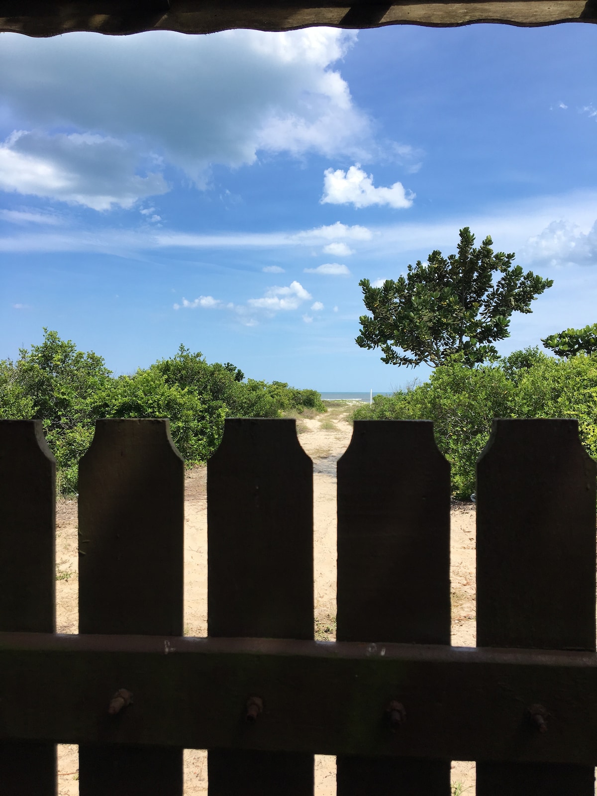 A view through a wooden fence opens up to a sandy pathway leading towards the beach. Lush greenery is visible on either side, with the ocean seen in the distance under a partially cloudy sky.