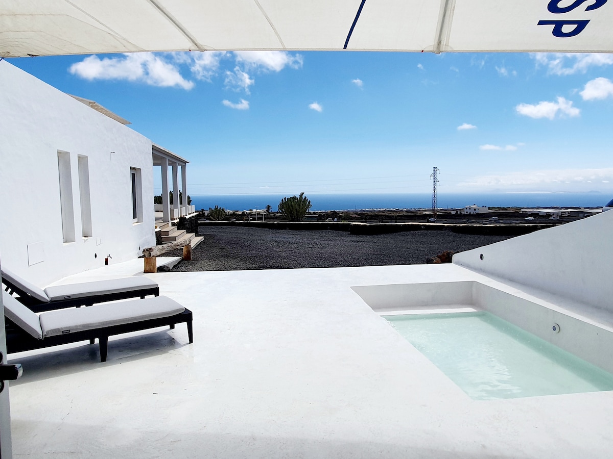 A sunlit terrace features comfortable loungers beside a small plunge pool. The expansive view includes the ocean under a clear blue sky, framed by minimalistic white architecture. Light-colored gravel surrounds the area, enhancing the sense of openness and tranquility.