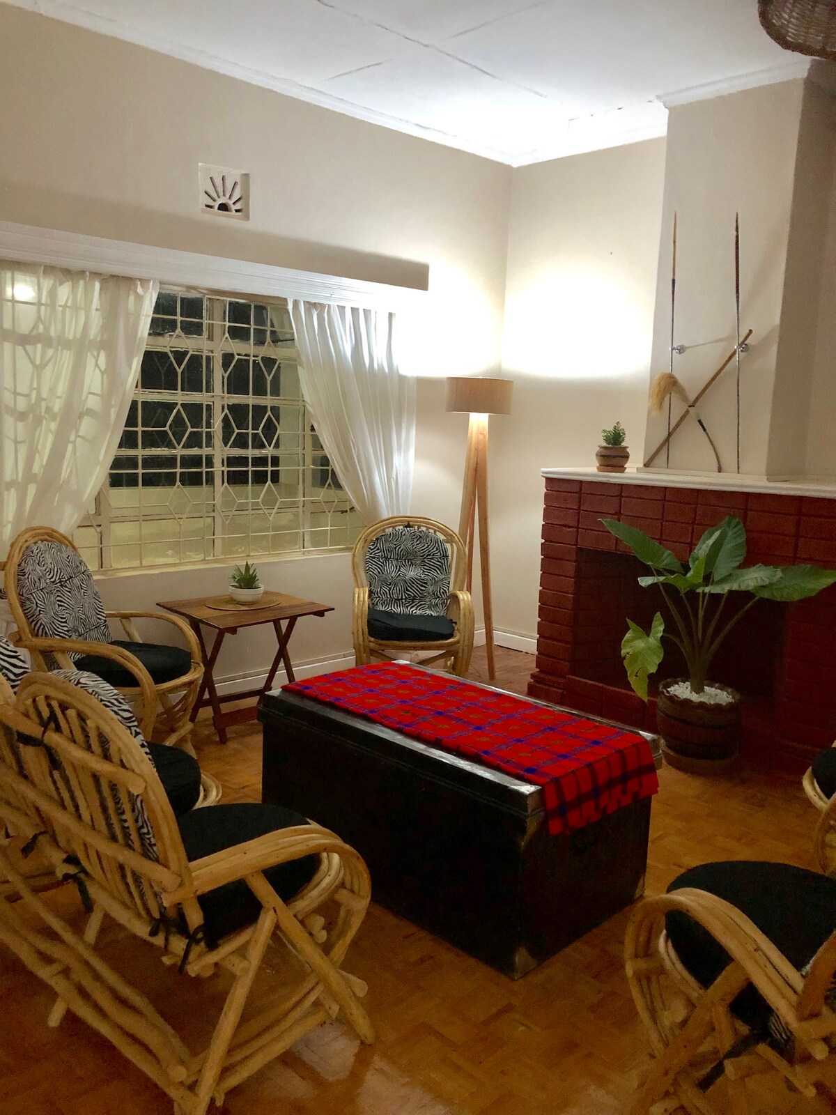A cozy living area features a central wooden table adorned with a vibrant red and blue blanket. Surrounding it are four rattan chairs and a pair of decorative plants. Soft lighting highlights the room's textures, while a large window ensures ample natural light.