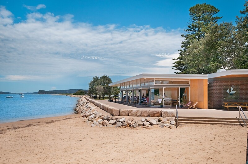 A modern waterfront building is positioned along a sandy beach, with large glass windows reflecting the surrounding landscape. A rocky shoreline separates the structure from the calm waters, and distant boats can be seen on the horizon under a partly cloudy sky.