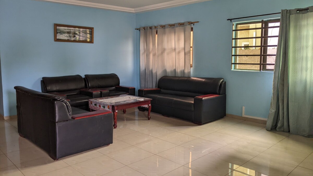 A spacious living area is furnished with comfortable black sofas and a central coffee table. Light beige tiles reflect natural light from the window, while soft blue walls create a calm ambiance. Sheer curtains allow light to filter through.