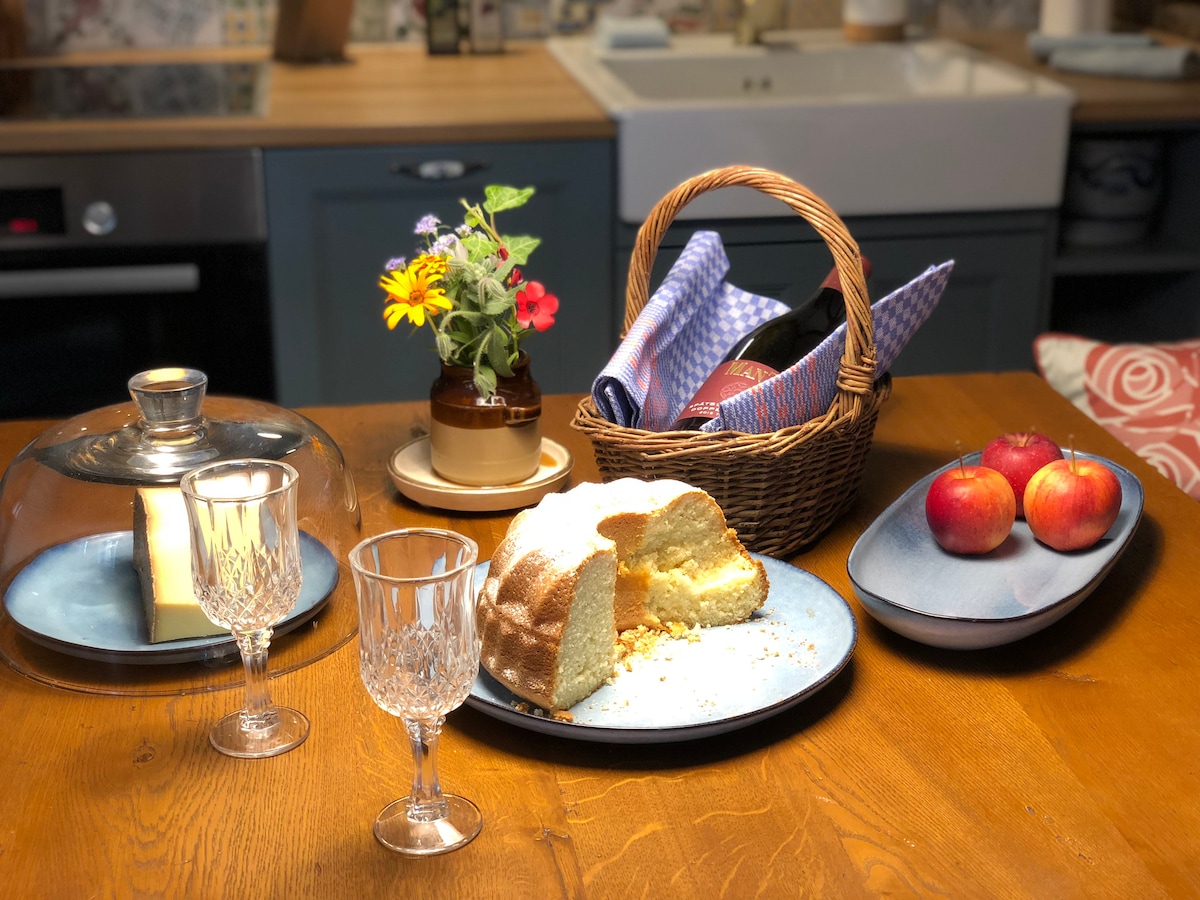 A wooden dining table is set with a variety of items. A glass cloche covers a slice of cake, while a basket includes wine and folded napkins. Two crystal glasses reflect light, and several fresh apples are displayed on a blue plate alongside vibrant flowers in a small pot.