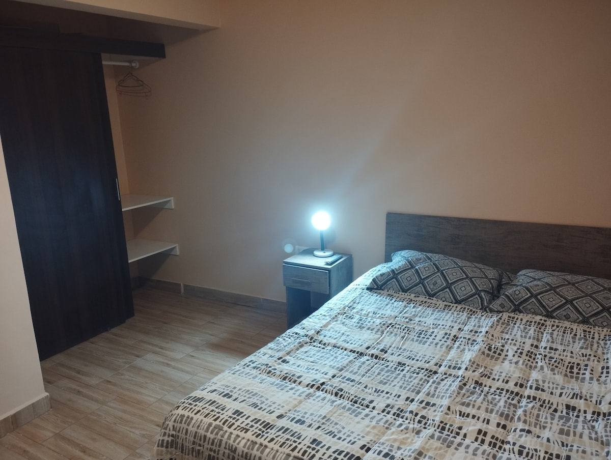 A well-lit bedroom features a spacious bed with a patterned comforter. A nightstand with a small lamp is positioned next to the bed. A closet with sliding doors offers additional storage, while a shelf is visible against the wall.