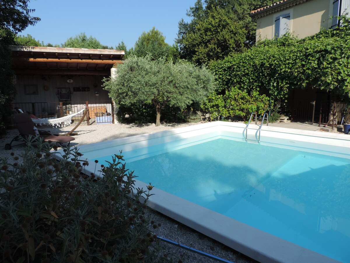 A private swimming pool is surrounded by lush greenery and gravel landscaping. Sun loungers are positioned beside the water, offering a place for relaxation. A shaded area with an outdoor structure can be seen in the background, providing additional seating and space for leisure.