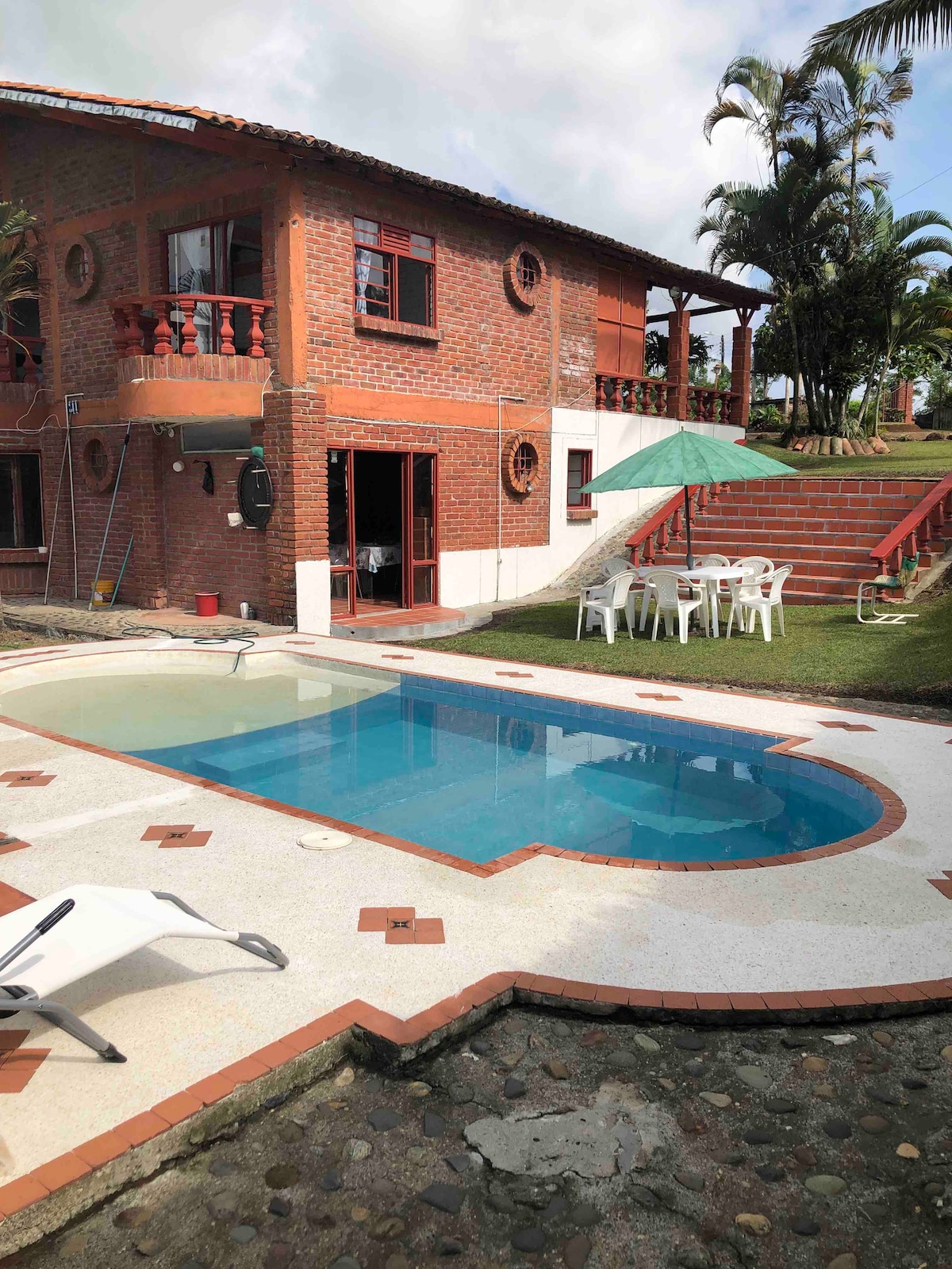A spacious pool area features a rectangular swimming pool surrounded by a tiled deck with lounge chairs. The red brick house with wooden balconies is visible in the background, along with a green umbrella over a table and chairs situated on the grassy area.