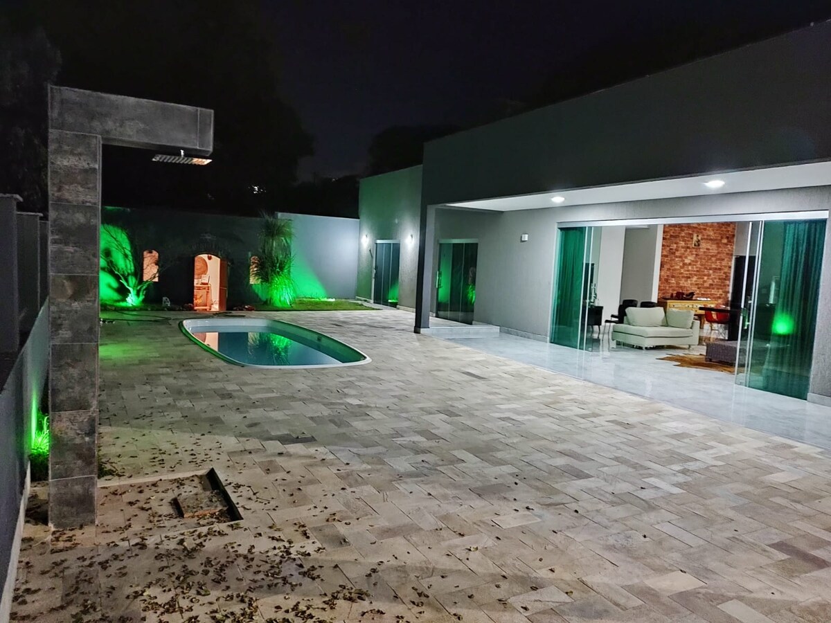 A modern outdoor space features a sleek pool, surrounded by stone tiles. Green lighting casts a soft glow on the pool area, enhancing the evening ambiance. Large glass doors provide a view into a cozy living area with a warm, brick wall.