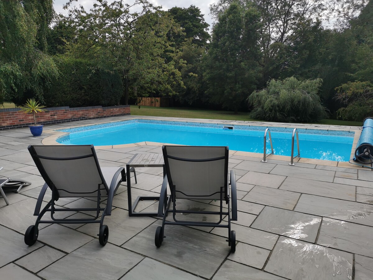 A well-maintained outdoor swimming pool is framed by a spacious slate patio. Two comfortable lounge chairs are positioned nearby, offering a relaxing spot to enjoy the surroundings. Lush greenery surrounds the area, providing a serene backdrop.
