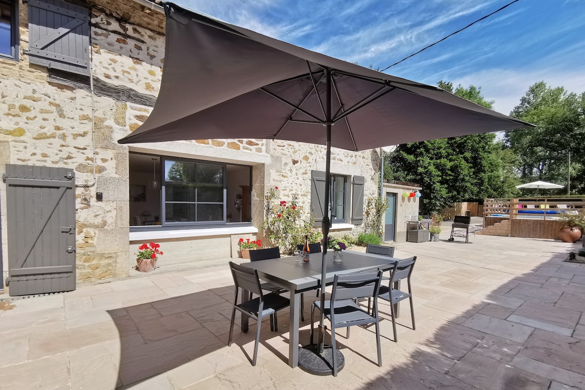 An outdoor dining area is featured with a sizable table surrounded by six chairs, shaded by a large umbrella. The rustic stone building provides a charming backdrop, while colorful flower pots add a touch of vibrancy to the inviting space.