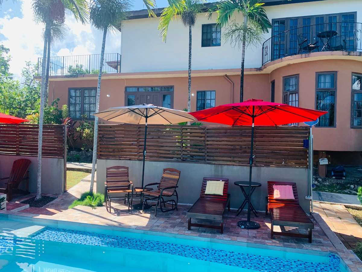 A shared pool area features two comfortable loungers beneath red umbrellas, positioned beside a cool blue water surface. In the background, the villa's exterior showcases a mix of windows and balconies surrounded by lush palm trees.