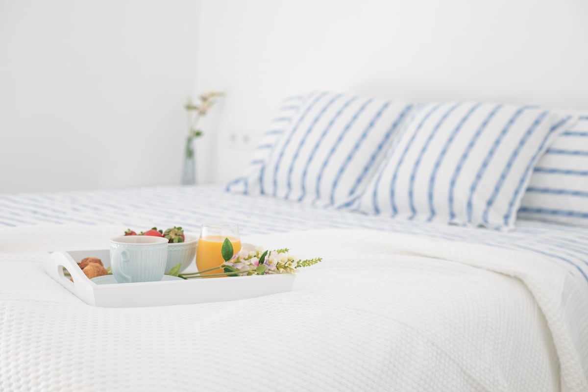 A spacious bed is adorned with white bedding featuring subtle textures. Striped pillows add a touch of colour. A tray rests on the bed, holding a bowl of fresh fruit, a croissant, and a glass of juice, complemented by delicate flower accents in the background.