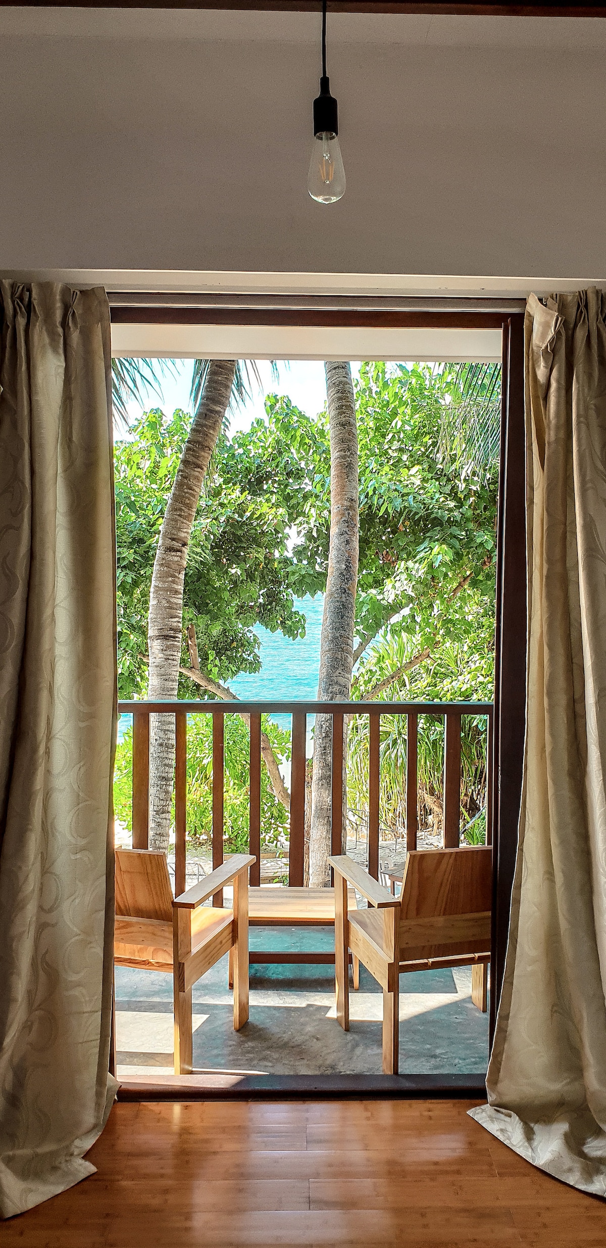 A serene view from inside the guest house features a private balcony with two wooden chairs. Lush tropical foliage and palm trees frame the view of the sparkling Indian Ocean visible through the open doorway, inviting relaxation and tranquility.