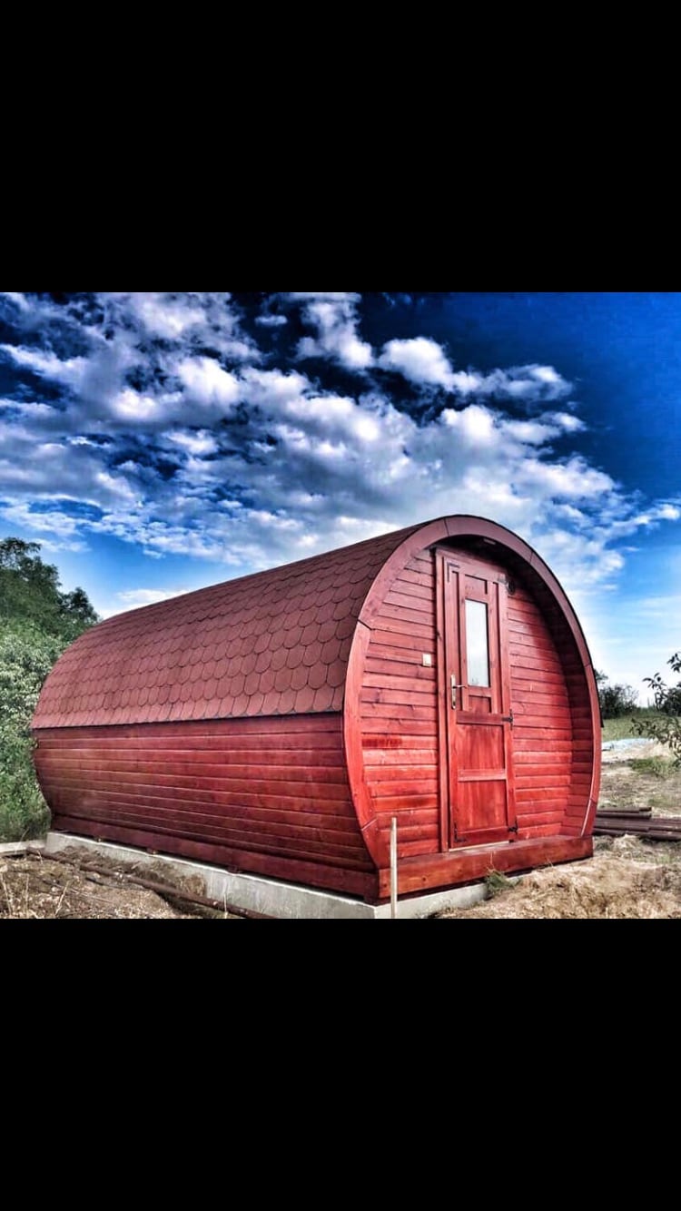 A rustic wooden barrel-shaped cabin with a sloped roof is showcased against a backdrop of a cloudy blue sky. The cabin features a small, paneled door framed by a light wooden border, set on a concrete base, blending harmoniously with the surrounding landscape.
