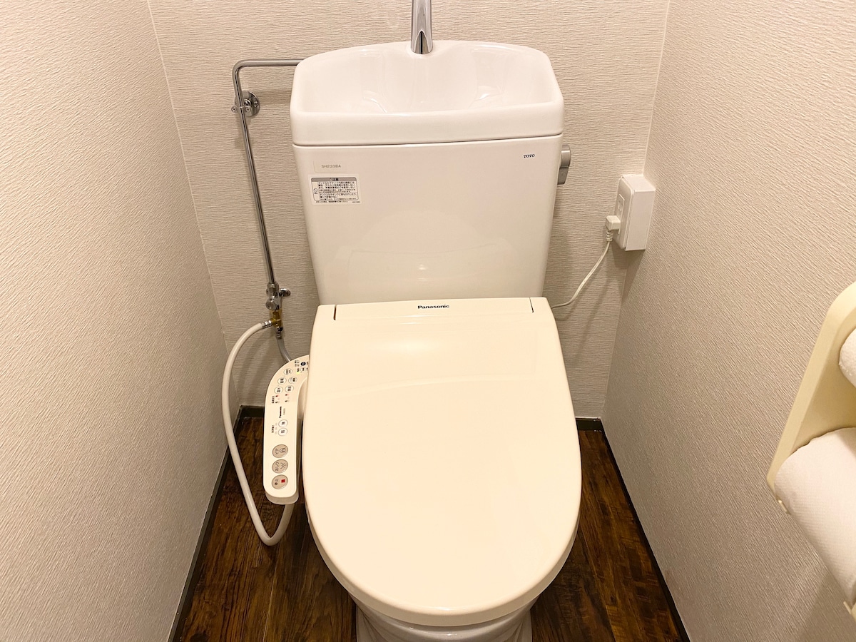 A modern toilet is positioned against a light-colored wall. Features include a sleek design with integrated bidet options. The flooring is made of dark wood, contrasting with the overall bright bathroom setting.