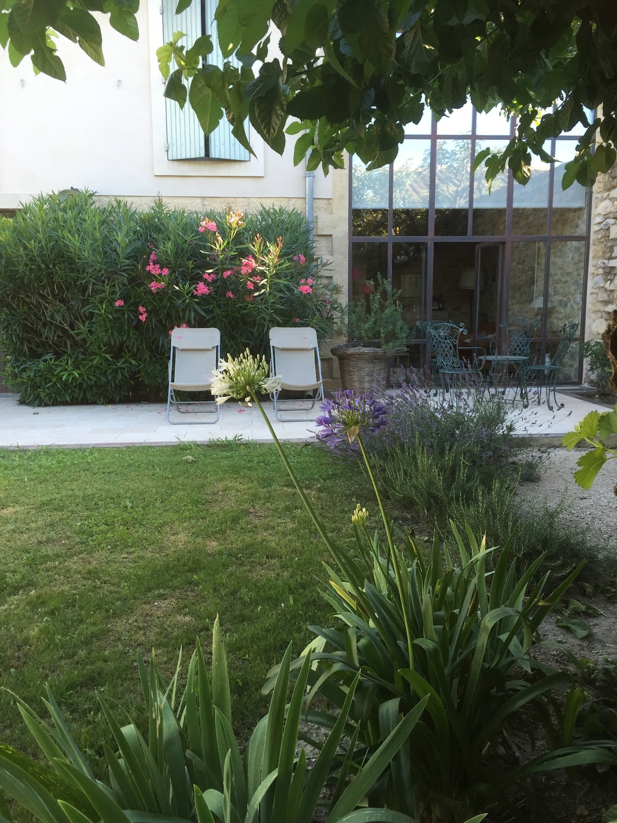 A charming garden area is presented, featuring well-maintained greenery and blooming flowers. Two lounge chairs are positioned on a stone pathway, with a dining set visible in the background. The façade of the house is partially seen through large windows, enhancing the tranquil outdoor space.